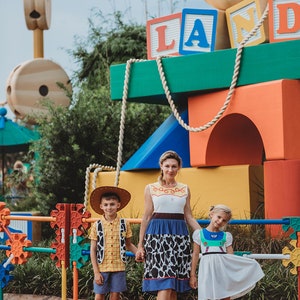 Puede incluir: Una familia de tres personas vestida como personajes de la pel&iacute;cula Toy Story. La madre lleva un vestido azul y blanco con una falda de estampado de vaca. El hijo lleva un chaleco amarillo y un sombrero de vaquero. La hija lleva un vestido blanco con un chaleco verde y azul. Est&aacute;n de pie frente a una gran estructura colorida con las palabras "Toy Story Land" en ella.