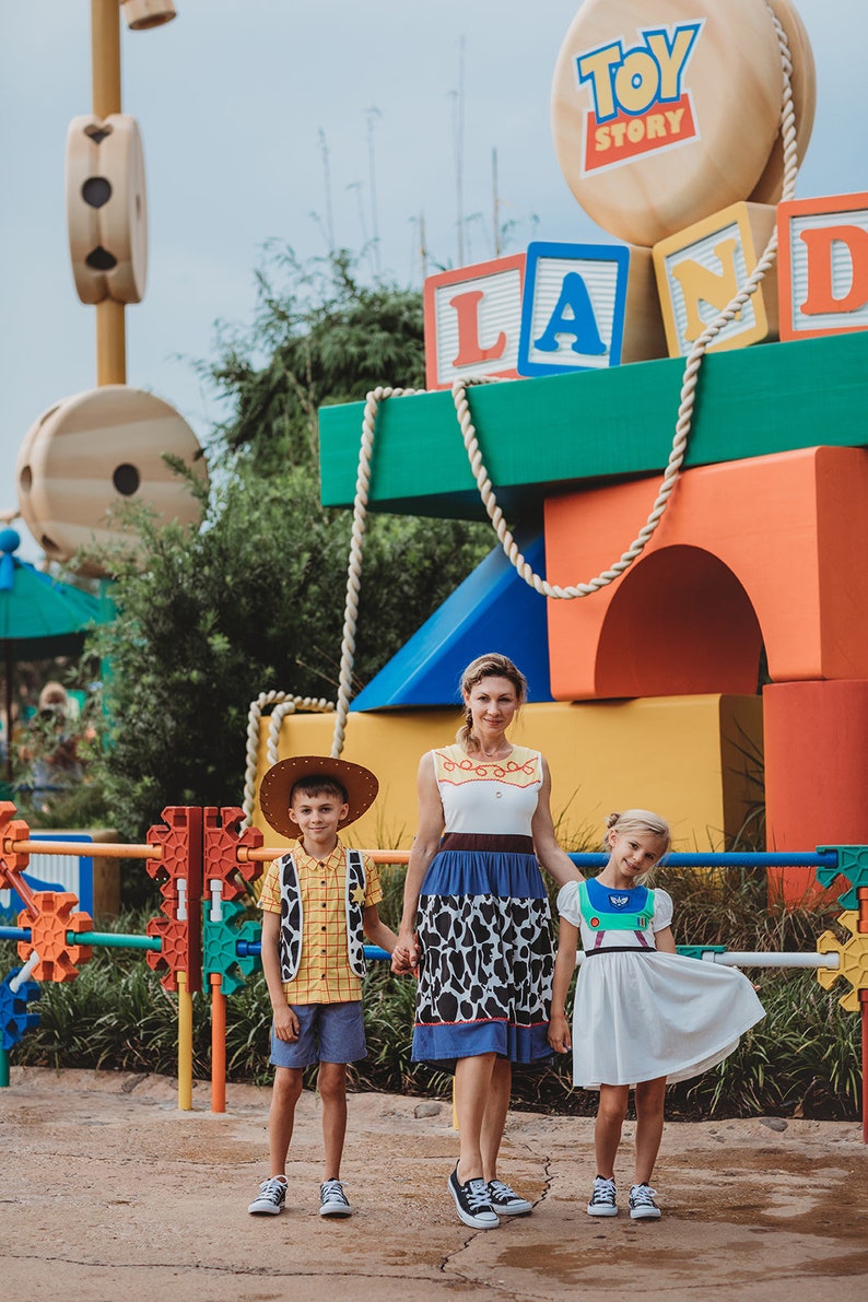 Puede incluir: Una familia de tres personas vestida como personajes de la pel&iacute;cula Toy Story. La madre lleva un vestido azul y blanco con una falda de estampado de vaca. El hijo lleva un chaleco amarillo y un sombrero de vaquero. La hija lleva un vestido blanco con un chaleco verde y azul. Est&aacute;n de pie frente a una gran estructura colorida con las palabras "Toy Story Land" en ella.