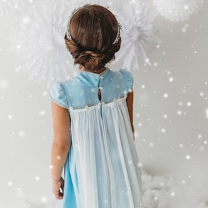 May include: A light blue dress with white accents and a high collar. The dress features a delicate floral pattern on the bodice and a flowing white skirt. The back of the dress has button closures. The girl's hair is styled in a bun with a silver headband.