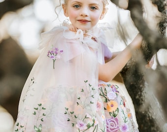Könnte beinhalten: Ein junger Mensch trägt einen floral bestickten Umhang und Rock, kombiniert mit einem Juwelen besetzten Diadem. Der Umhang und der Rock weisen zarte Blumenstickereien in Lila, Rosa und Gelb auf. Das Diadem ist silberfarben mit rosa und blauen Juwelen.