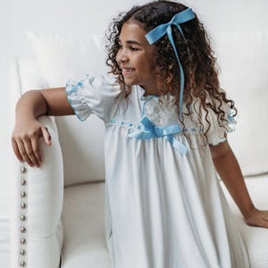 May include: A white nightgown with light blue trim and a matching bow. The short-sleeved dress features a round neckline and a bow at the chest. The girl's hair is styled with a light blue bow.