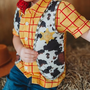 May include: A child's cowboy costume featuring a yellow plaid shirt with red lines, a cow-print vest, and a sheriff's badge. The outfit includes a red bandana and is paired with blue jeans. The costume is designed for a playful, Western-themed look.