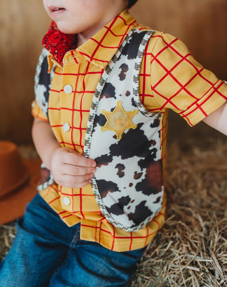 May include: A child's cowboy costume featuring a yellow plaid shirt with red lines, a cow-print vest, and a sheriff's badge. The outfit includes a red bandana and is paired with blue jeans. The costume is designed for a playful, Western-themed look.