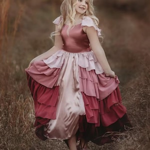 A girl is wearing the Yule Love all of the ruffles Maxi Dress in shades of pink and red. There are pink flutter sleeves. The bodice is a pink mauve and leads into cascading layers chiffon that start with dusty pink and flow down into a deep maroon.