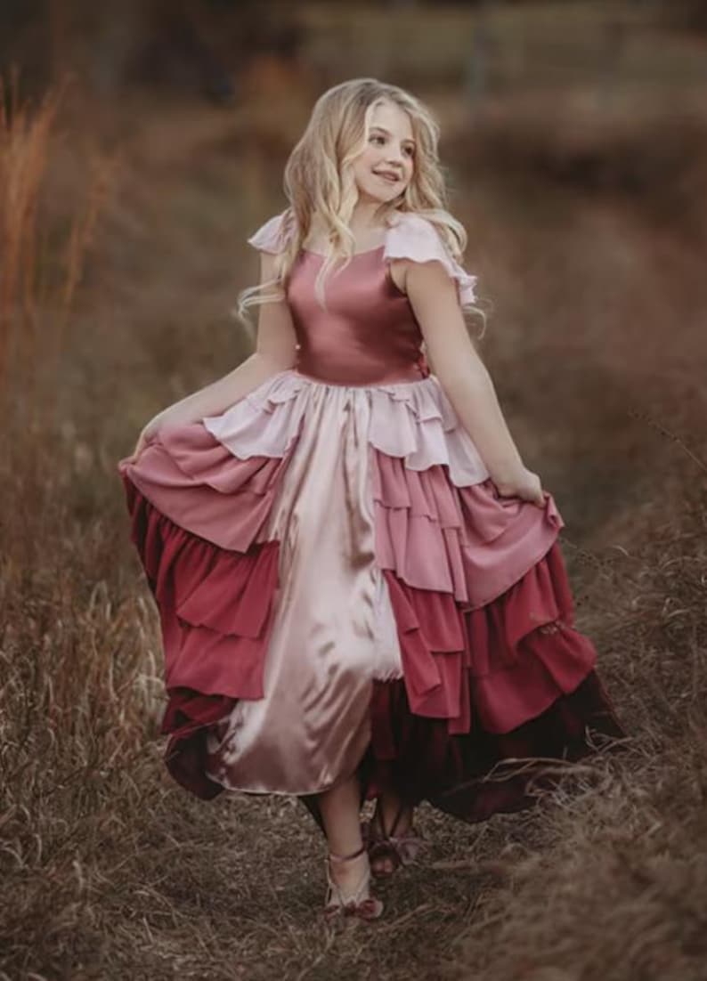 A girl is wearing the Yule Love all of the ruffles Maxi Dress in shades of pink and red. There are pink flutter sleeves. The bodice is a pink mauve and leads into cascading layers chiffon that start with dusty pink and flow down into a deep maroon.