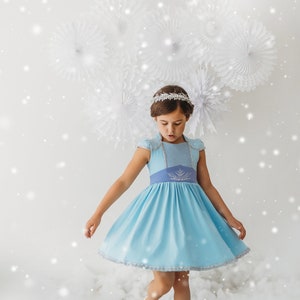 May include: A light blue dress with a silver trim and a matching blue sash. The dress has short sleeves and a decorative design on the bodice. A silver tiara is worn on the head. The background features white paper decorations and a snowy effect.