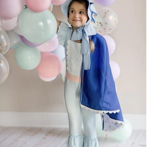 May include: A young girl wearing a light blue dress with a white lace overlay and a blue cape. She is wearing a light blue bonnet with a white lace trim. She is standing in front of a wall of pastel balloons.