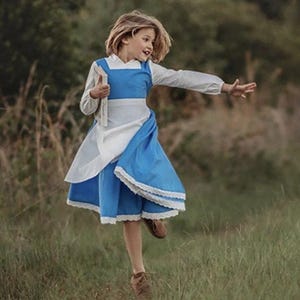 A young girl stands wearing the Red Rose Princess Village Dress from the Fairy Godmother Collection, reminiscent of Disney's Belle from "Beauty and the Beast." The white sleeves are long. The dress has a bight blue chest and skirt.