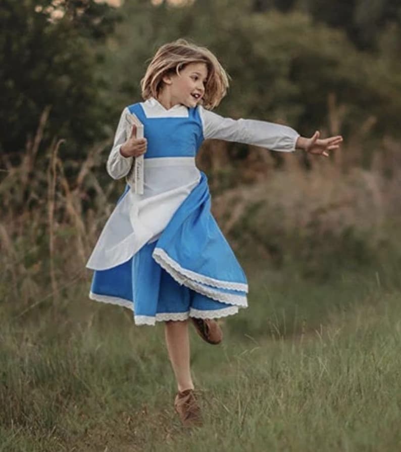 A young girl stands wearing the Red Rose Princess Village Dress from the Fairy Godmother Collection, reminiscent of Disney's Belle from "Beauty and the Beast." The white sleeves are long. The dress has a bight blue chest and skirt.