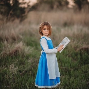 A young girl stands wearing the Red Rose Princess Village Dress from the Fairy Godmother Collection, reminiscent of Disney's Belle from "Beauty and the Beast." The white sleeves are long. The dress has a bight blue chest and skirt.