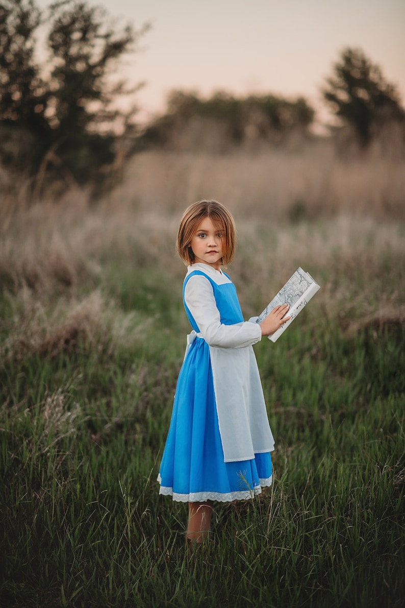 A young girl stands wearing the Red Rose Princess Village Dress from the Fairy Godmother Collection, reminiscent of Disney's Belle from "Beauty and the Beast." The white sleeves are long. The dress has a bight blue chest and skirt.