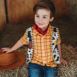 May include: A child's cowboy costume featuring a yellow plaid shirt with red lines, a cow-print vest, a red bandana, blue jeans, and a brown cowboy hat. The outfit is designed to resemble a character from a popular animated film.