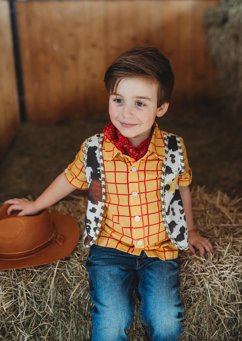 May include: A child's cowboy costume featuring a yellow plaid shirt with red lines, a cow-print vest, a red bandana, blue jeans, and a brown cowboy hat. The outfit is designed to resemble a character from a popular animated film.