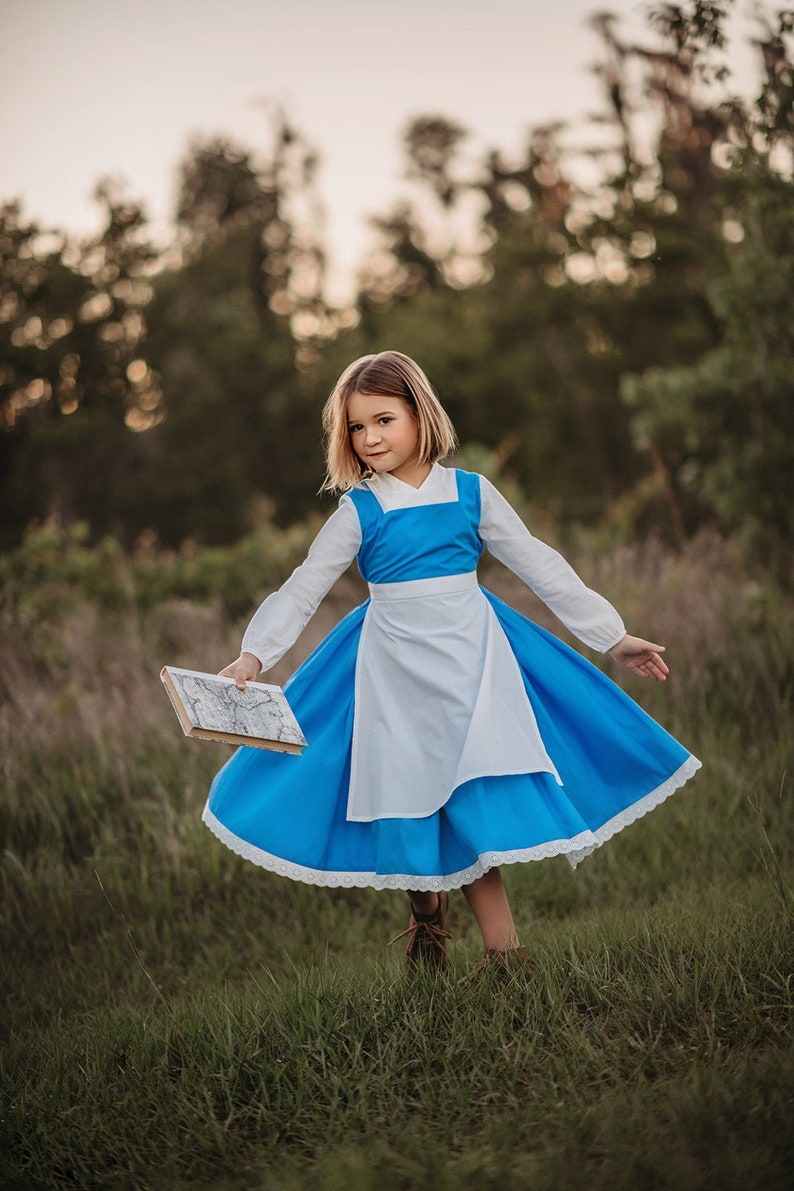 A young girl stands wearing the Red Rose Princess Village Dress from the Fairy Godmother Collection, reminiscent of Disney's Belle from "Beauty and the Beast." The white sleeves are long. The dress has a bight blue chest and skirt.