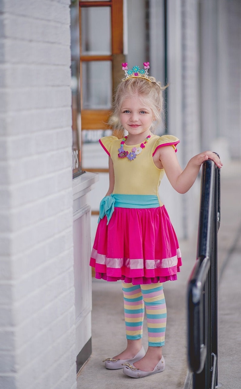 Pu&ograve; includere: Una bambina che indossa un vestito giallo e rosa con un nastro turchese e una corona di fiori. Indossa anche leggings a righe colorate e scarpe bianche.