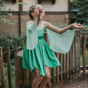 Pu&ograve; includere: Una ragazza indossa un vestito verde con maniche fluttuanti e un corpetto bianco. L'abito ha una gonna svasata ed &egrave; abbinato a scarpe bianche. La ragazza &egrave; in piedi su un ceppo d'albero vicino a una staccionata di legno.