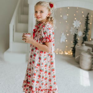 Könnte beinhalten: Ein junges Mädchen trägt ein langes Kleid mit Blumenmuster und roten Schleifen und Rüschen. Das Kleid hat einen weißen Hintergrund mit roten und blauen floralen Akzenten. Sie hält einen kleinen, runden Behälter und trägt braune Hausschuhe. Der Hintergrund umfasst eine weiße Treppe und dekorative Elemente.