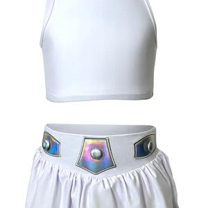 Puede incluir: Conjunto blanco de dos piezas con top corto de cuello alto y falda. La falda tiene un dobladillo con volantes y un cinturón decorativo con detalles iridiscentes y tachuelas plateadas. El conjunto está hecho de una tela lisa, probablemente sintética.