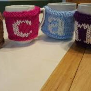 Puede incluir: Cuatro tazas de cerámica blanca con cubiertas de punto. Cada cubierta es de un color diferente: marrón, rosa, azul y morado. Las cubiertas tienen letras blancas que deletrean la palabra "COLD".