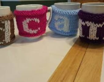 May include: Four white ceramic mugs with knitted cosy covers. Each cosy is a different colour: brown, pink, blue, and purple. The cozies have white letters on them spelling out the word "COLD".