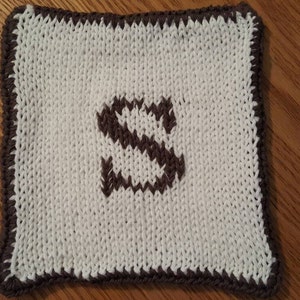 May include: A white knitted square with a brown knitted letter "S" in the center. The square has a brown knitted border.