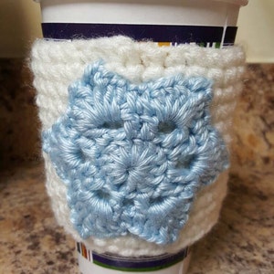 Könnte beinhalten: Weiße gehäkelte Kaffeetassen-Hülle mit einem hellblauen Schneeflocken-Design.
