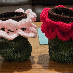 May include: Four crocheted flower pot covers in different colors: yellow, pink, red, and white. Each cover has a green base and a crocheted flower design on top.