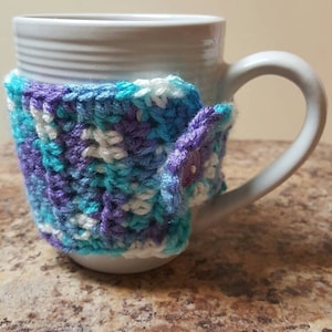 Könnte beinhalten: Eine weiße Keramiktasse mit einer gehäkelten Hülle. Die Hülle besteht aus einer Mischung aus blauem, lila und weißem Garn, das um die Tasse gewickelt ist. Die Tasse steht auf einer braunen, gesprenkelten Oberfläche.