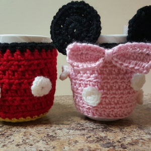 Könnte beinhalten: Zwei gehäkelte Kaffeetassen-Cozies, einer rot mit schwarzen Ohren und weißen Punkten, der andere rosa mit schwarzen Ohren und einer weißen Schleife.