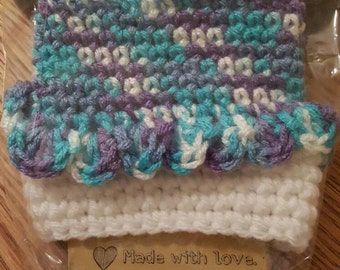 May include: A crocheted cup cosy in shades of purple, teal, and white with a scalloped edge. The cosy is packaged in a clear plastic bag with a brown card that reads "Cup Cozy" and "Made with love."