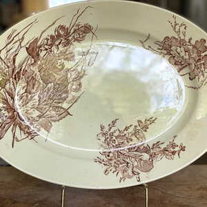 May include: An oval-shaped, white ceramic serving platter with a brown floral design. The design features stylized leaves and flowers.