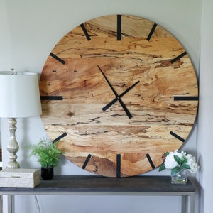 May include: A large wooden clock with black hands and black Roman numeral markings on a light wood background. The clock is hanging on a white wall above a gray console table with a woven basket and a white throw.