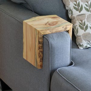 May include: A wooden side table with a natural wood finish and live edge design. The table is attached to the arm of a gray sofa.