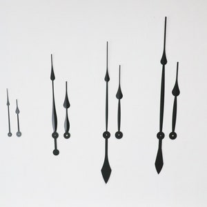 May include: Black clock hands of various sizes and styles are arranged against a white background. The hands have different shapes, including pointed and rounded tips, and are designed for clock making or repair.
