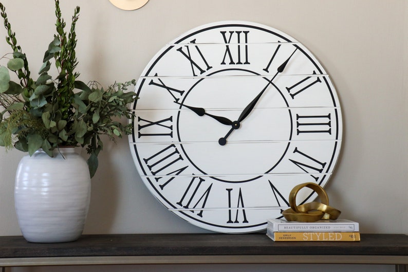 May include: A large white farmhouse style clock with black Roman numeral markings and black hands. The clock is hanging on a wall above a dark brown console table with a white vase filled with greenery and a gold metal sculpture on top of two books.