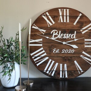 Puede incluir: Un reloj de madera grande con n&uacute;meros romanos y la palabra "Blessed" en escritura blanca. El reloj tambi&eacute;n tiene el texto "Est. 2020" debajo de la palabra "Blessed".