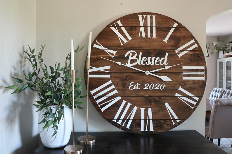 Puede incluir: Un reloj de madera grande con n&uacute;meros romanos y la palabra "Blessed" en escritura blanca. El reloj tambi&eacute;n tiene el texto "Est. 2020" debajo de la palabra "Blessed".