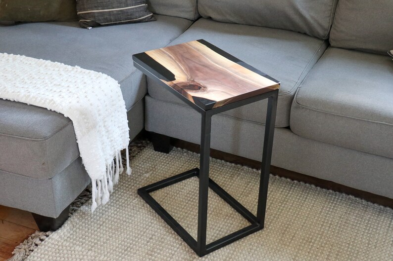 May include: A black metal side table with a live edge walnut wood top and black epoxy resin inlay. The table is sitting on a light beige rug in front of a gray couch.