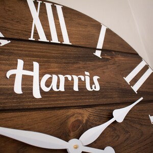 Puede incluir: Un reloj de madera marr&oacute;n con marcas de n&uacute;meros romanos blancos y manecillas blancas. La esfera del reloj tambi&eacute;n tiene el nombre "Harris" escrito en letras blancas cursivas.