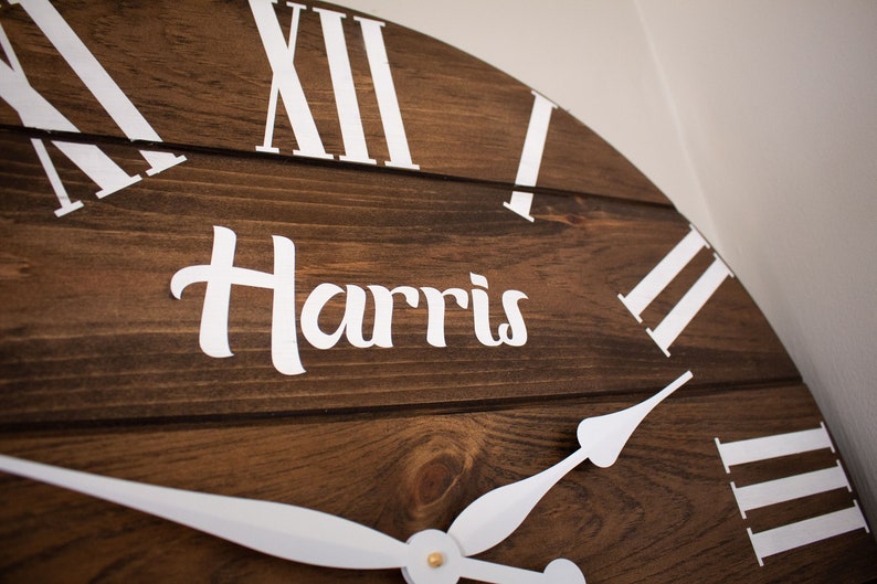 Puede incluir: Un reloj de madera marr&oacute;n con marcas de n&uacute;meros romanos blancos y manecillas blancas. La esfera del reloj tambi&eacute;n tiene el nombre "Harris" escrito en letras blancas cursivas.