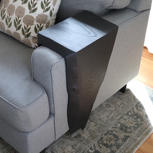 May include: A black wooden side table attached to the arm of a gray couch. The table has a unique, angled design and is perfect for holding a drink or snack while relaxing on the couch.