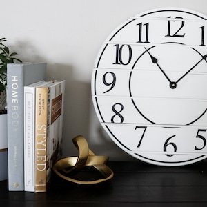 May include: A white clock with black numbers and hands, a potted plant, and three books titled "Homebody", "Beautifully Organized", and "Styled" on a black surface.