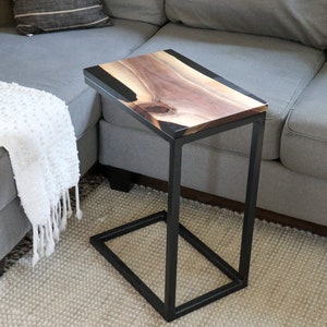 May include: A black metal side table with a live edge walnut wood top and black epoxy resin inlay. The table is sitting on a light gray rug in front of a gray couch.