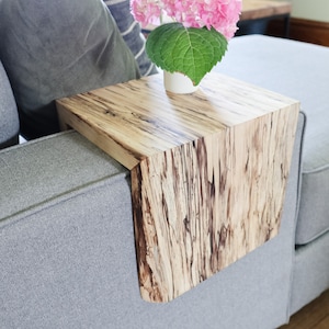 May include: A wooden side table with a natural wood grain finish. The table is attached to the arm of a gray couch and has a rounded edge. A vase with pink flowers sits on the table.