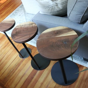 May include: Three modern side tables with round, dark wood tops and black metal bases. The wood grain is visible on the table tops. The tables are different heights and are set on a hardwood floor.