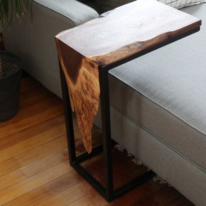 May include: A wooden side table with a live edge top and a black metal frame. The table is placed next to a gray couch.