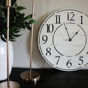 May include: A white farmhouse style clock with black numbers and hands. The clock is hanging on a wall with two gold candle holders on a black surface.