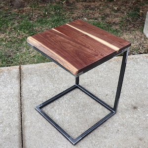 May include: A small, square, wooden table with a live edge top and a black metal frame. The table is sitting on a concrete surface.