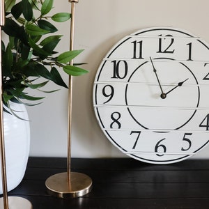 May include: A white farmhouse style clock with black numbers and hands. The clock is hanging on a wall with two gold candle holders and a white vase with green plants.