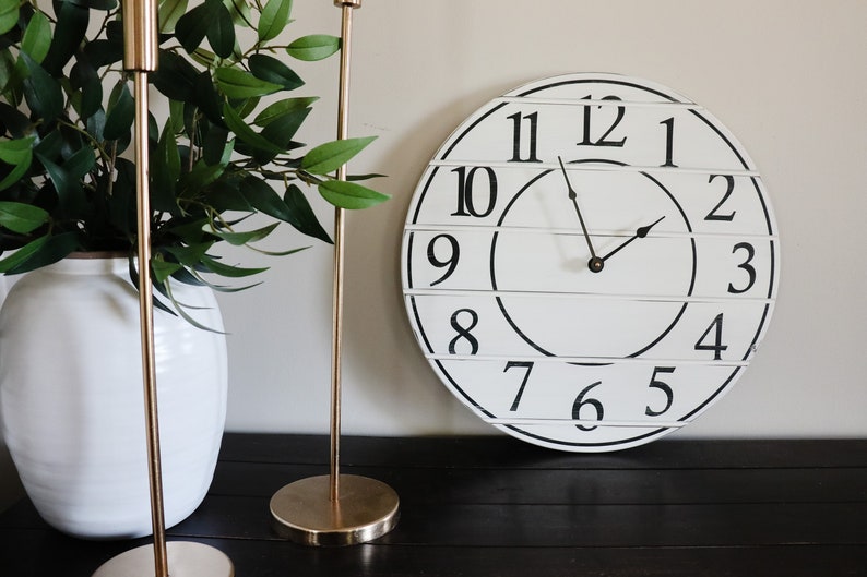 May include: A white farmhouse style clock with black numbers and hands. The clock is hanging on a wall with two gold candle holders and a white vase with green plants.
